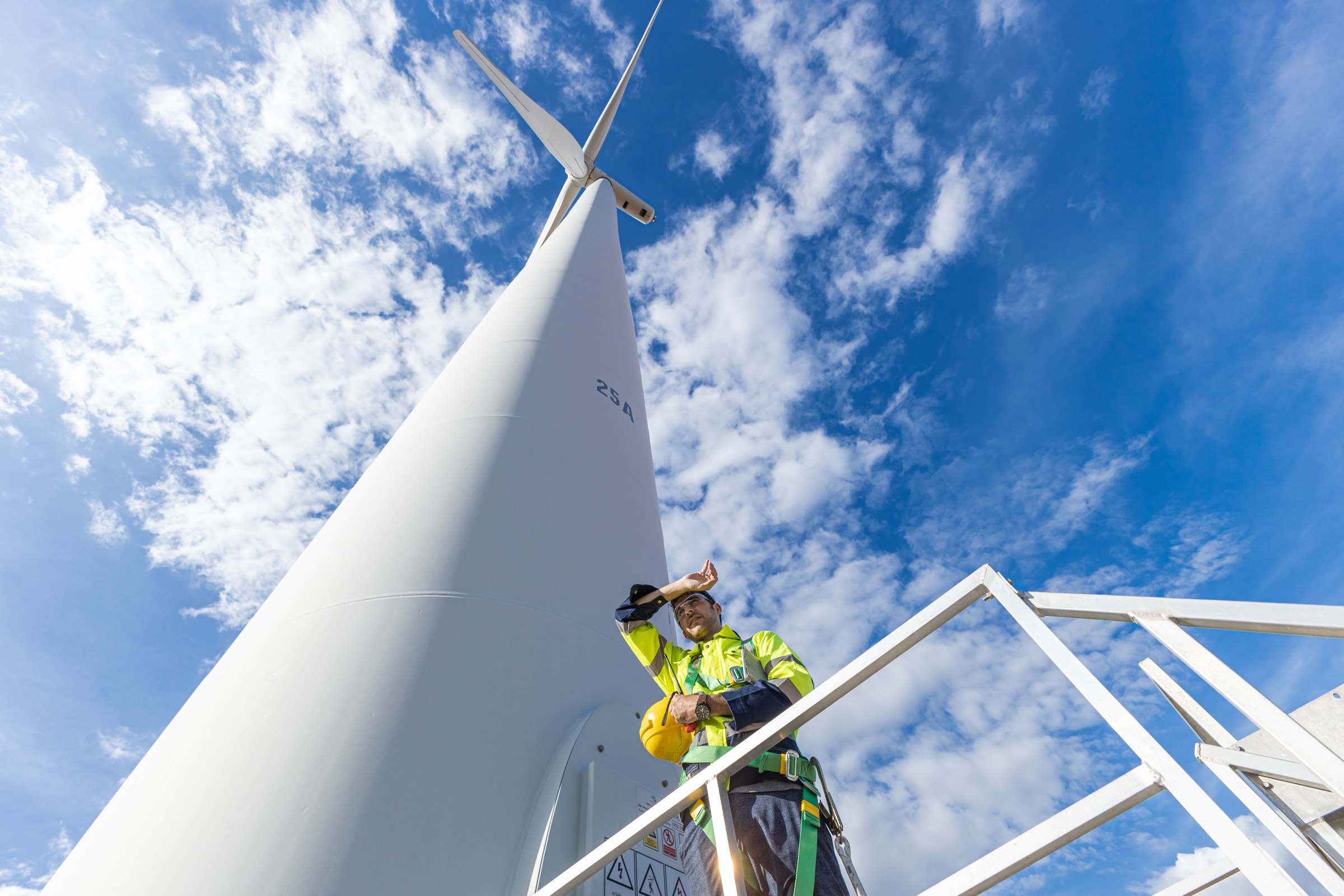 A wind turbine technician standing on a platform high on a turbine tower against a bright sky.