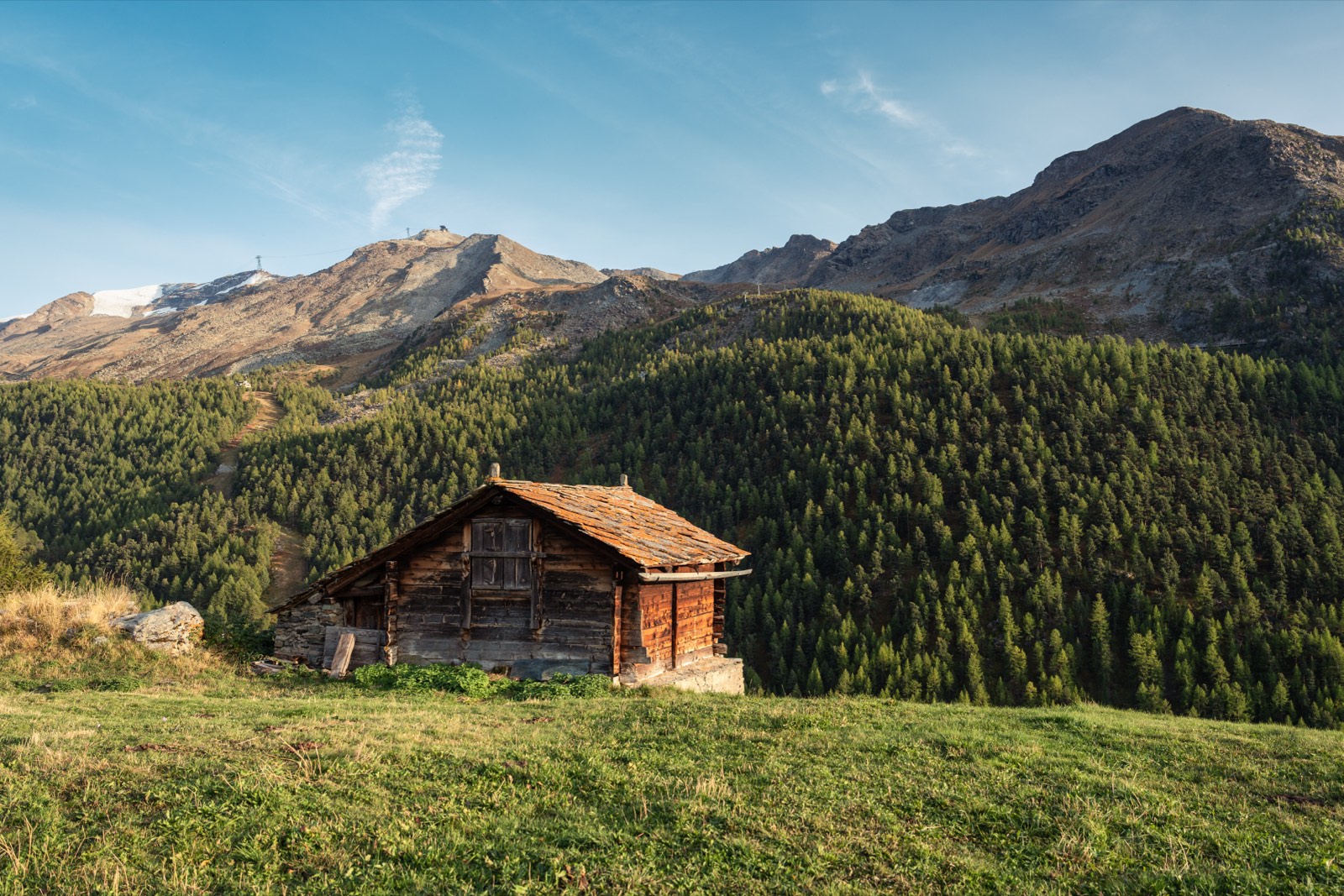 A weathered timber cabin on a grassy slope below forested mountains.