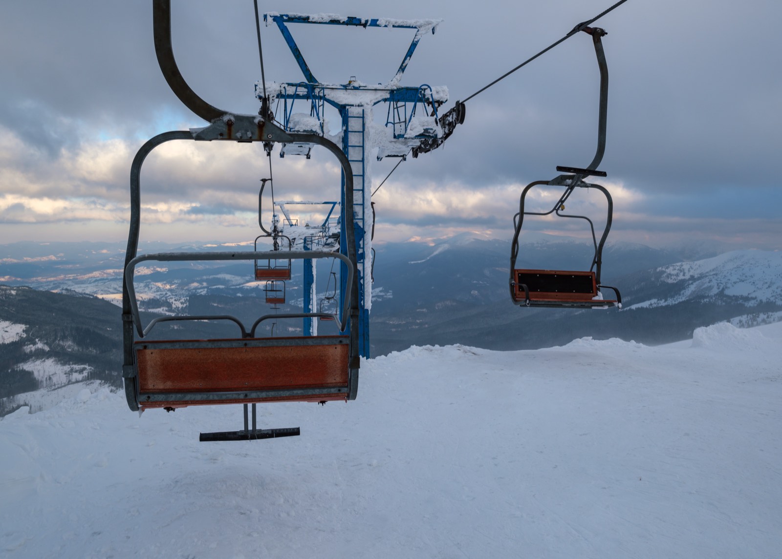Empty chairlift chairs above a snow-covered piste with mountain ridges and heavy cloud behind.