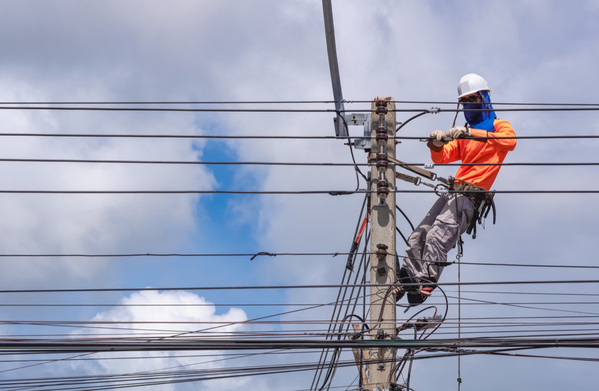 A power-line worker in safety gear working on overhead cables at the top of a utility pole.